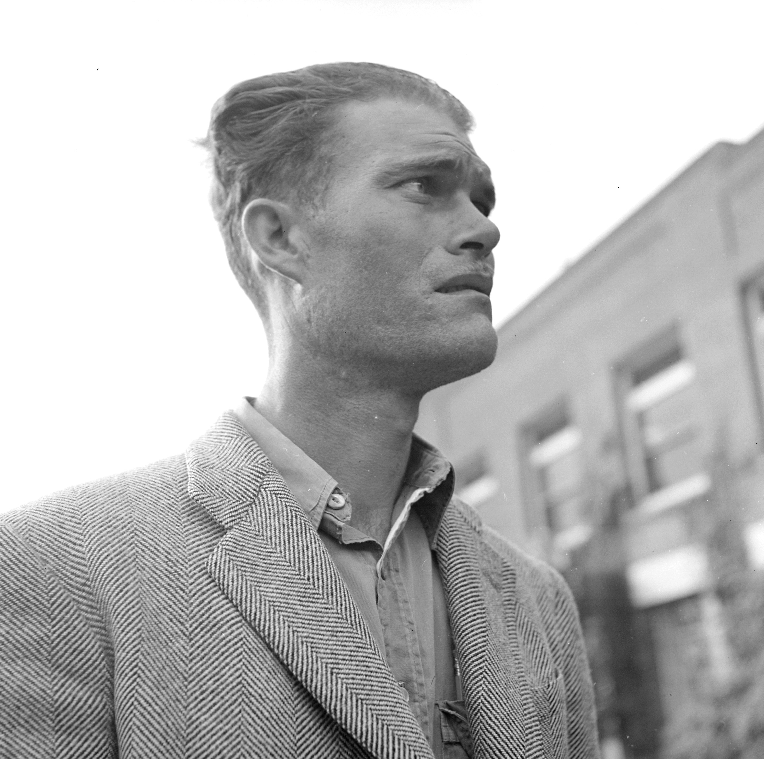 A black-and-white photograph of a young man in a tweed jacket looking upward with an expression of anticipation and anxiety, seated outdoors near a brick building