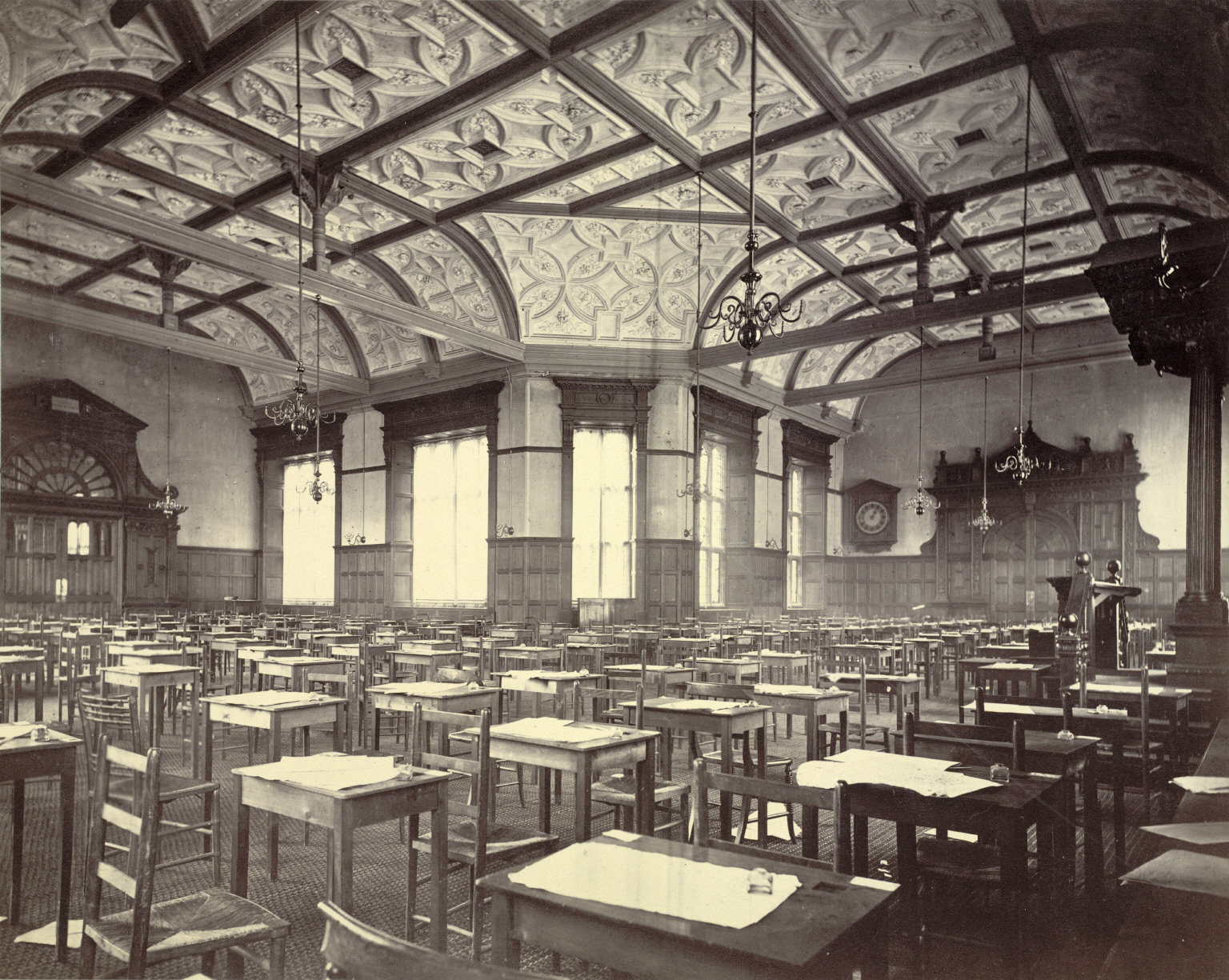A vast, ornate Victorian-era examination hall with rows of empty wooden desks, each set with paper and inkwell, illuminated by tall curtained windows and gas-style chandeliers